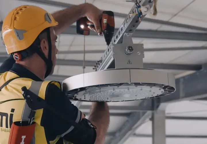 Pracownik w żółtym kasku i kamizelce montuje nowoczesne oświetlenie LED. To wymiana oświetlenia na LED – remont czy modernizacja hali.