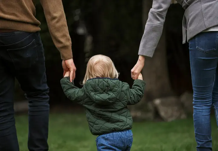 Dziecko z blond włosami, w zielonej kurtce, trzyma za ręce rodziców. To obraz przysposobienia niepełne, pełnego miłości i nadziei.