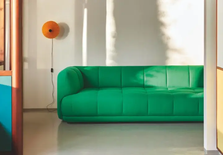 Ein leuchtend grünes Sofa steht vor einer weißen Wand, an der eine orangefarbene Lampe hängt. Sonnenlicht wirft Schatten. Mette hay.