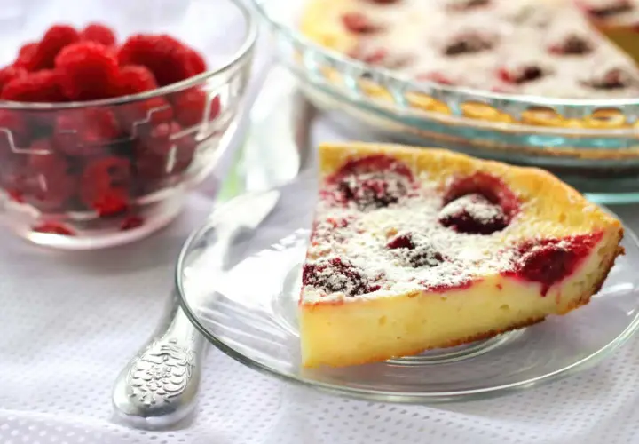 Une part de clafoutis framboise, saupoudrée de sucre glace, à côté d'un bol de framboises fraîches.