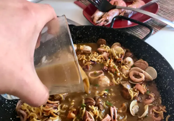 Vertiendo caldo en una paellera con fideos, calamares y almejas. Así se hace fideuá.