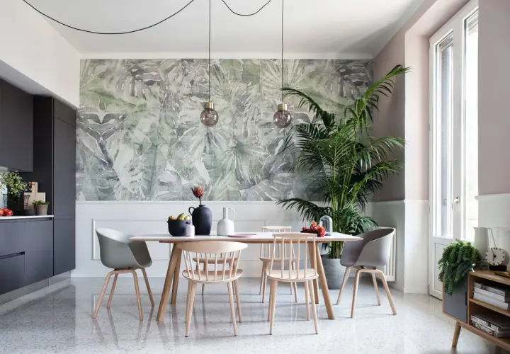 Un soggiorno moderno con carta da parati a tema tropicale, un tavolo da pranzo con sedie in legno e un'elegante cucina grigia. Chi mette la carta da parati sceglie un tocco di natura.