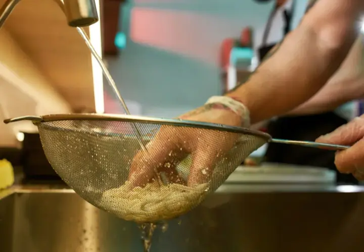 Ręce w rękawiczkach płuczą makaron pod bieżącą wodą, by zapobiec jego sklejaniu.