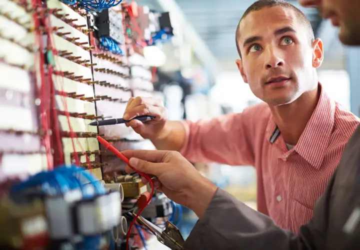 Dwóch mężczyzn pracuje przy panelu elektrycznym, jeden trzyma miernik.