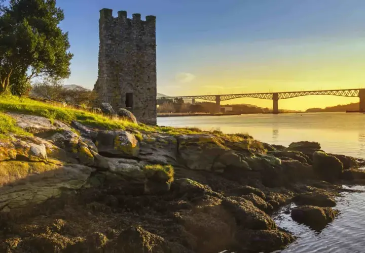 Torre medieval junto a un río al atardecer, con un puente al fondo. Imagina el tren Madrid Ferrol pasando por allí.