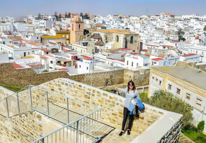 Una mujer sonríe en un balcón de piedra con vistas a un pueblo blanco. ¡Descubre todo **que ver en Tarifa**!