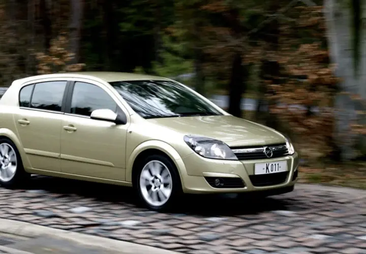 Złoty Opel Astra H 1.6 benzyna w ruchu na brukowanej drodze, otoczony jesiennym lasem.