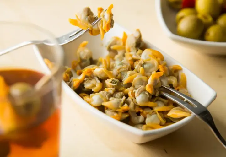 Un plato de berberechos, pequeños moluscos marinos, listos para disfrutar. Se ven deliciosos junto a un vaso de bebida y aceitunas.