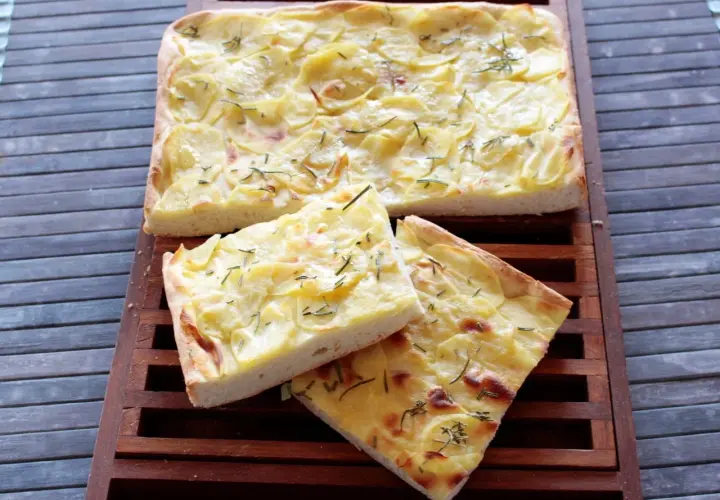Focaccia con lievito istantaneo, ricoperta di patate sottili e rosmarino, tagliata a fette su un tagliere di legno.