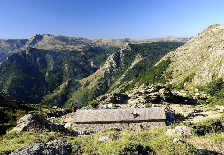 Kamienny schron na GR20 Korsyka, otoczony skalistymi zboczami i zielonymi dolinami.