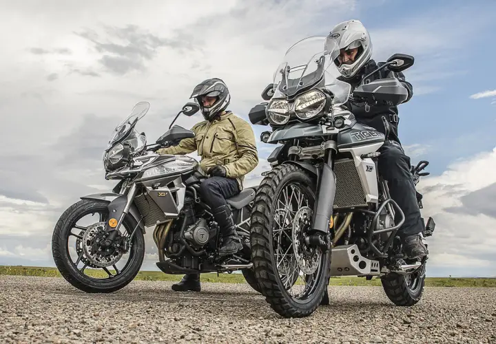 Dos motociclistas con cascos y equipo de aventura listos para explorar caminos y **trail asfalticas** bajo un cielo nublado.