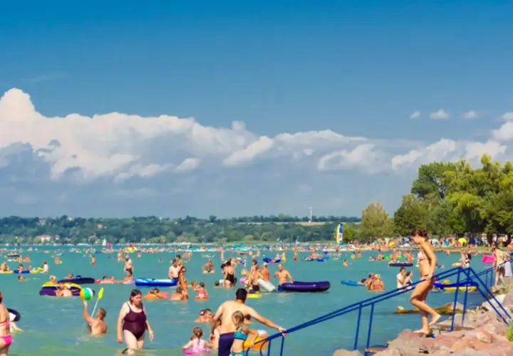 Tłum ludzi cieszy się latem na **jeziorze Balaton**. Woda pełna jest pływających, a na brzegu widać drzewa i skaliste nabrzeże.