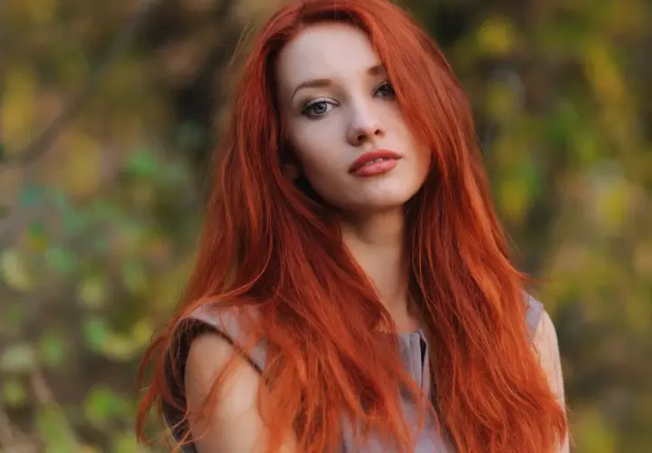 Eine Frau mit langen, dunkelroten Haaren blickt nachdenklich in die Kamera. Ihr Haar fällt ihr über die Schultern, während sie im Freien steht.