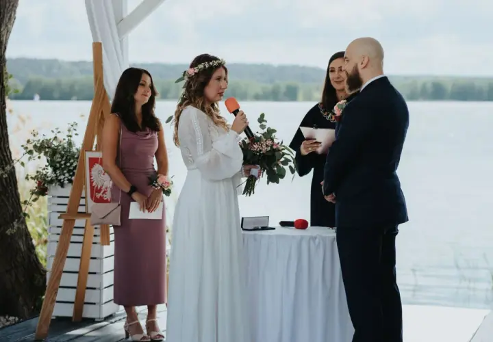 Panna młoda z mikrofonem i bukietem mówi podczas ceremonii. Obok stoi pan młody, urzędniczka i świadkowa. To ślub jednostronny nad jeziorem.