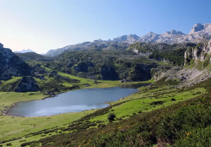 Lago en un valle montañoso verde con picos rocosos al fondo.
