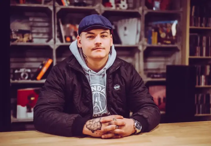 Młody mężczyzna w czapce i kurtce, z tatuażami na rękach, siedzi przy stole. W tle regały z płytami i innymi przedmiotami. To jego **frosti wiek**.