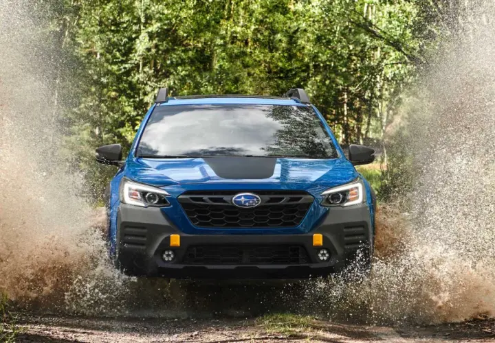 Niebieski Subaru Outback Wilderness z impetem wjeżdża w kałużę, tworząc fontannę wody i błota.