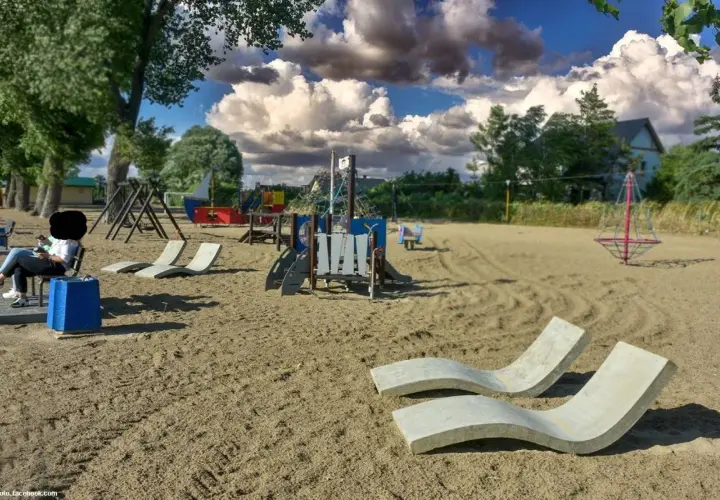 Pusta lubczyna plaża z placem zabaw i leżakami. Kobieta siedzi na ławce, patrząc na telefon.