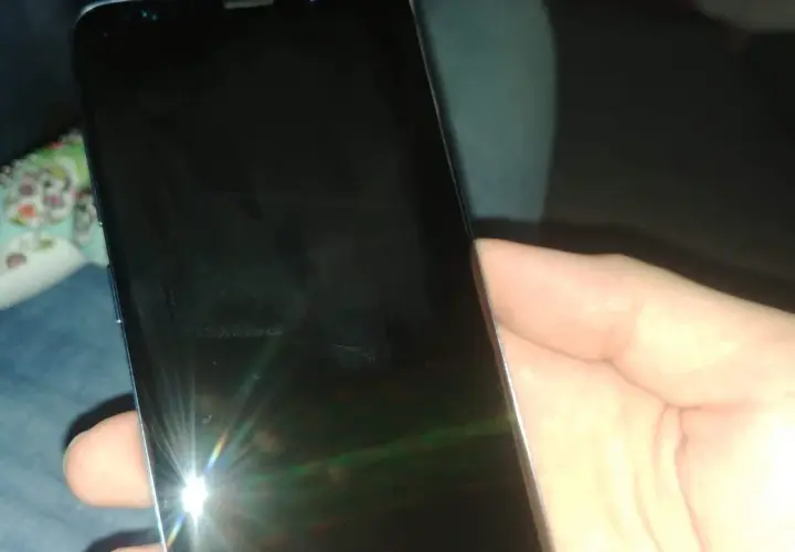 Dłoń trzyma czarny telefon, na którego ekranie widać odbicie światła i ledwo widoczną kreskę na telefonie.