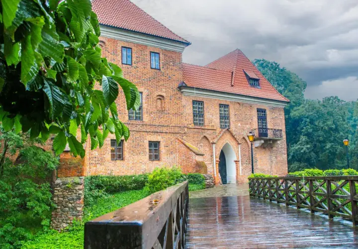 Deszczowy dzień nad zamkiem w Oporowie. Drewniany mostek prowadzi do ceglanego budynku, otoczonego zielenią.