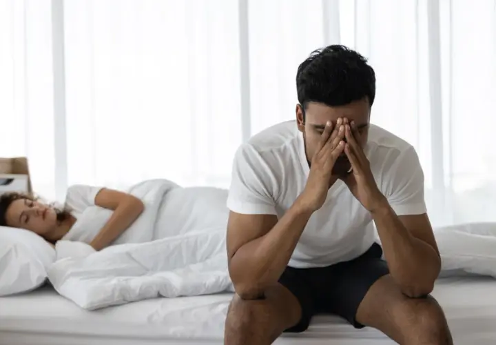 Uomo seduto sul letto con le mani sul viso, preoccupato per l'eiaculazione precoce. La donna dorme nel letto alle sue spalle.
