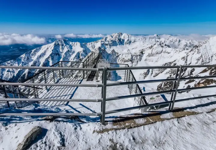 Widok z tarasu widokowego na szczycie Łomnicy. Stalowe balustrady i schody prowadzą do zapierającej dech w piersiach panoramy ośnieżonych gór.