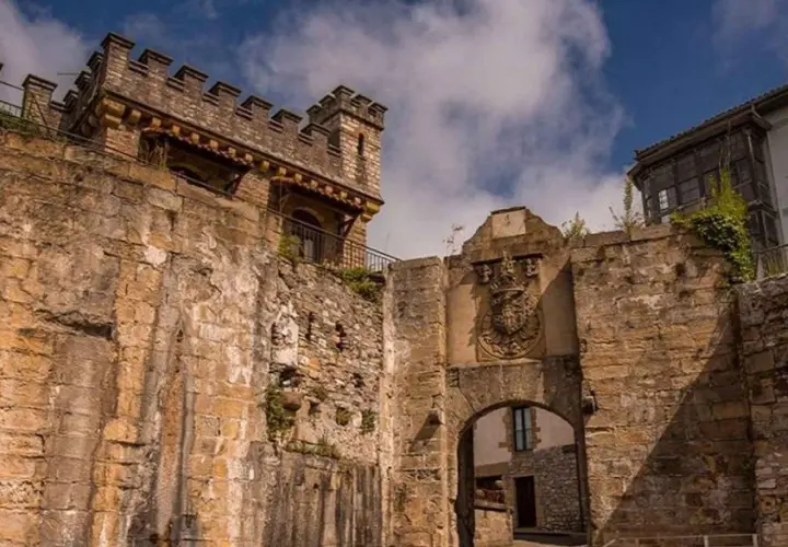 Antigua muralla y arco de entrada en Fuenterrabía, con un escudo nobiliario y un cielo azul con nubes.