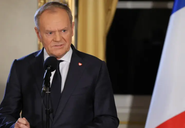 Donald Tusk w garniturze przemawia przy mikrofonie, obok polska flaga.
