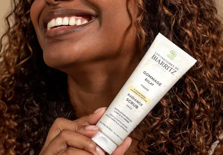 Une femme souriante tient un tube de gommage visage éclat. Ce produit sert à lisser et illuminer la peau, révélant sa beauté naturelle.