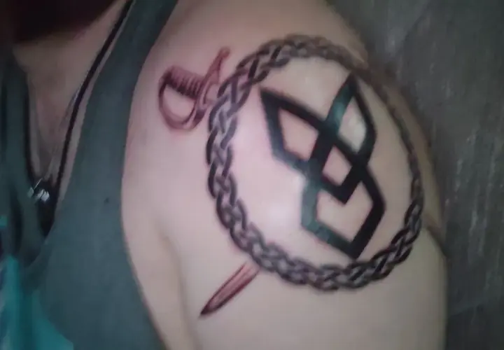 Tatuaż wojownik na ramieniu: spleciony łańcuch, szabla i symbol.