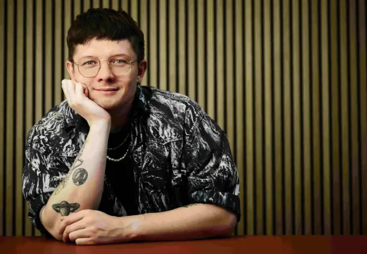 Fritz Espenlaub, mit Brille und Tattoos, lehnt nachdenklich am Tisch vor einer vertikalen Wand.