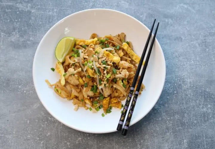 Pad thai z kurczakiem, jajkiem, orzeszkami i limonką na białym talerzu z pałeczkami.