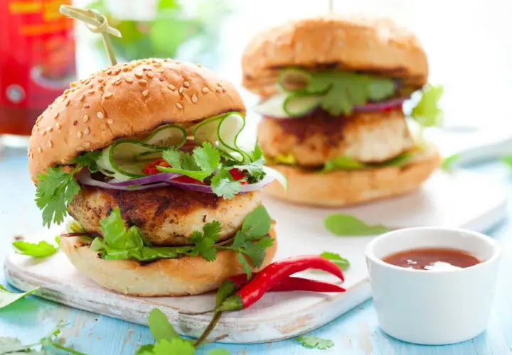 Dos hamburguesas de pollo frescas y jugosas, adornadas con pepino, cebolla morada, cilantro y chile rojo.