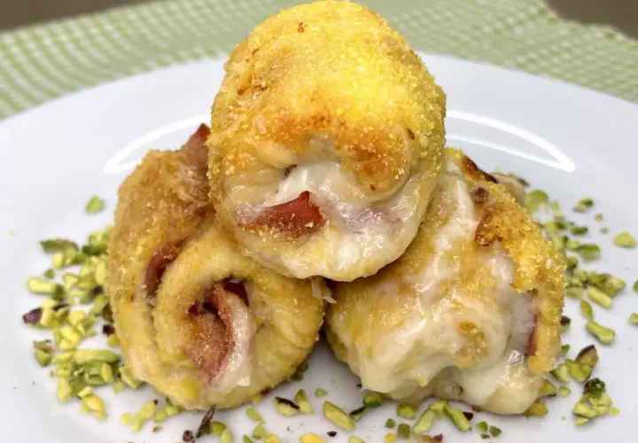 Tre involtini di pollo dorati e ripieni di formaggio filante e prosciutto, adagiati su un letto di pistacchi tritati.