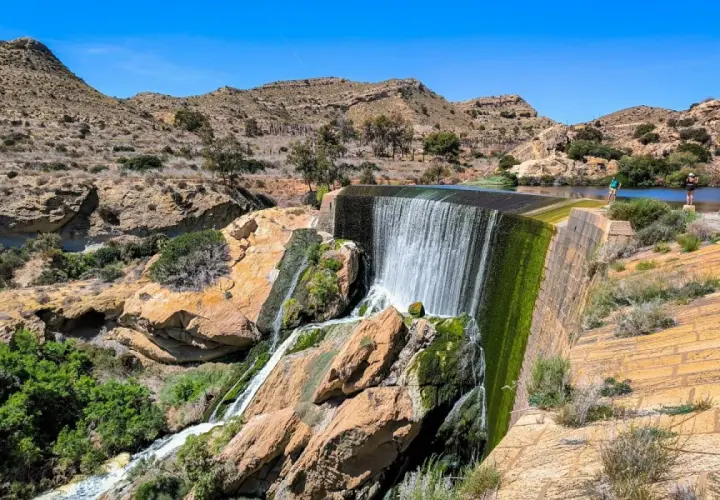 Cascada en el pantano de Elche, con rocas cubiertas de musgo y vegetación verde. Personas observan el agua.