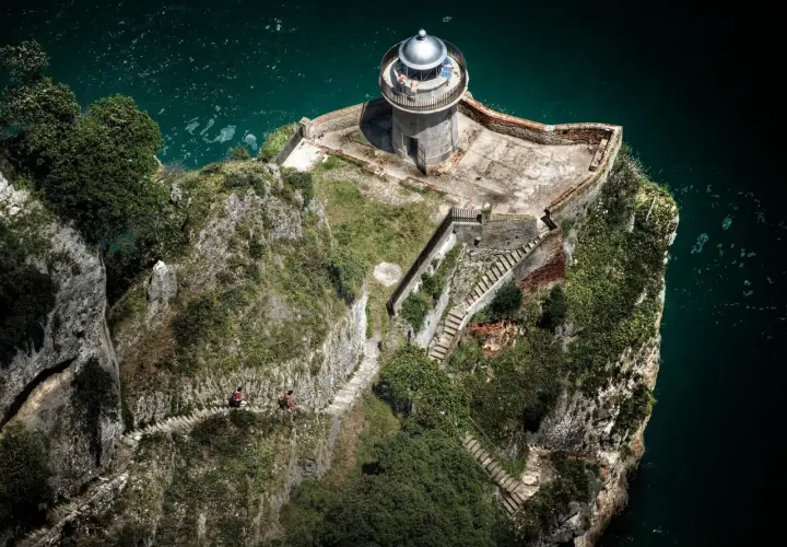Dos personas caminan por un sendero empinado hacia el faro de caballo, un faro solitario en un acantilado rocoso sobre el mar turquesa.