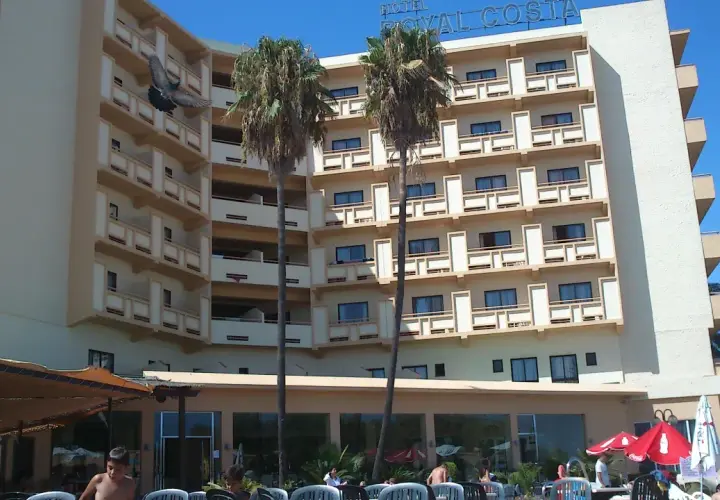Paloma vuela frente al Hotel Royal Costa, un edificio de varios pisos con balcones y palmeras.