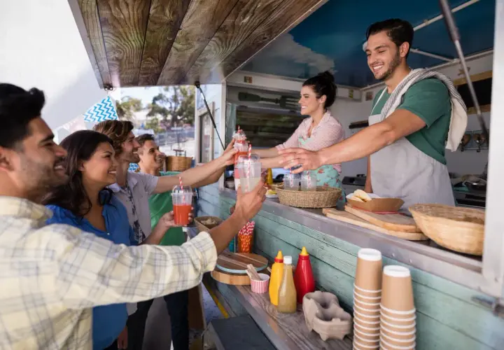 Uśmiechnięci klienci odbierają napoje od barmana w food trucku. Ciekawe, ile zarabia barman za noc przy takiej popularności!