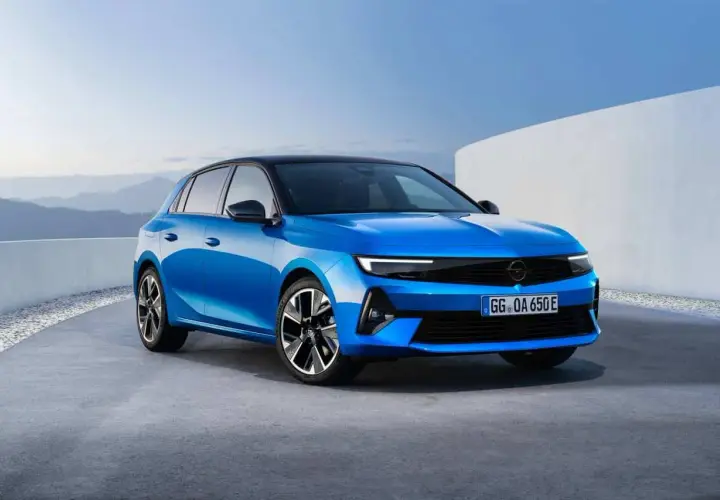 Błękitny opel elektryczny z czarnym dachem, zaparkowany na betonowej nawierzchni, z górzystym krajobrazem w tle.