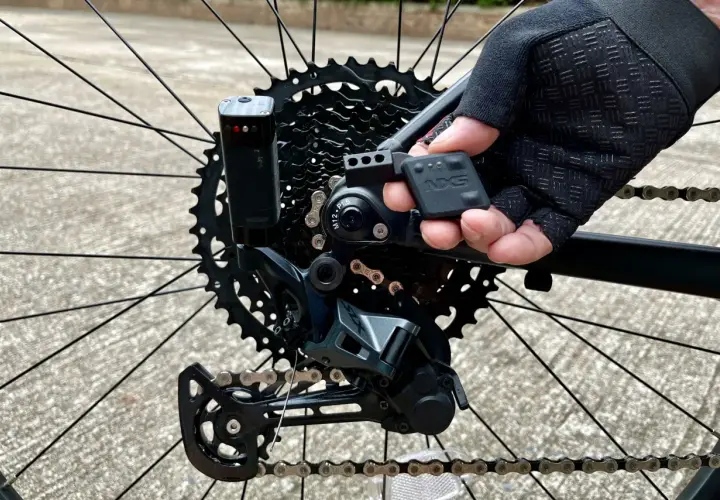 Close-up of a mountain bike's rear derailleur, showing the NX5 electronic shifting system being adjusted by a gloved hand.