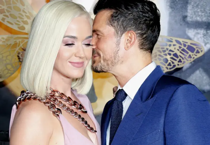 Orlando Bloom szepcze coś do ucha Katy Perry, która ma na sobie różową sukienkę i gruby, złoty naszyjnik.