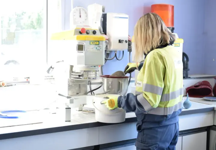 Trabajadora en laboratorio, usando guantes, manipula un recipiente con material. Se investiga qué es la silicona y sus usos en la industria.