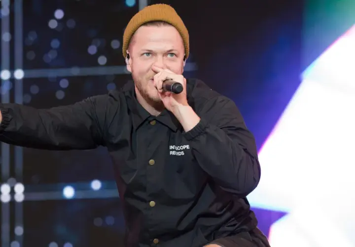 Imagine Dragons wokalista, Dan Reynolds, w beżowej czapce i czarnej kurtce, śpiewa do mikrofonu, pokazując znak pokoju.