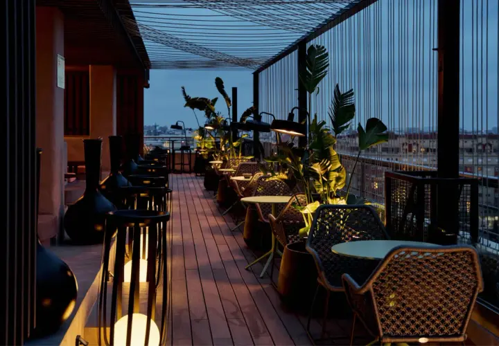 Terraza del Novotel Valencia con mesas y sillas de mimbre, iluminada por lámparas y rodeada de vegetación.