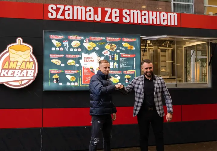 Dwóch mężczyzn wita się przy budce z kebabem, na której widnieje menu.