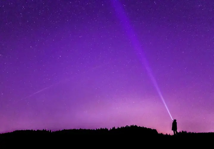 Sylwetka człowieka oświetla latarką nocne niebo, na którym widać gwiazdy i **gwiazdozbiór Tarczy**.