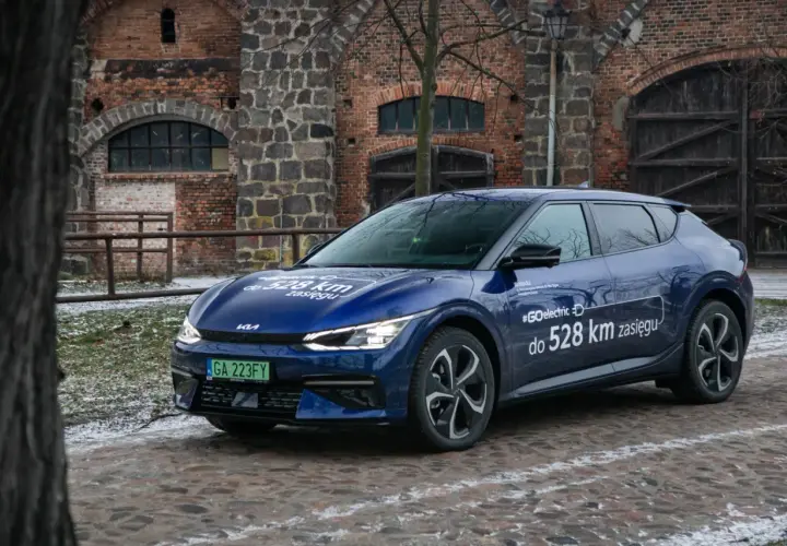 Niebieska Kia EV6 z napisem "#GOelectric D do 528 km zasięgu" stoi przed zabytkową ceglaną budowlą.