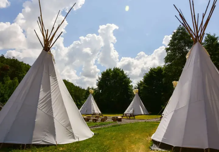 Malowniczy **pole namiotowe** z białymi tipi, drewnianymi ławami i stołami, otoczone zielenią i błękitnym niebem z chmurami.