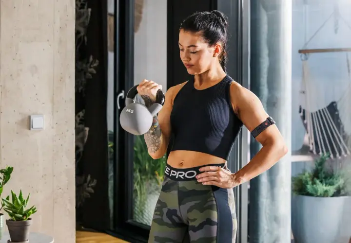 Kobieta ćwiczy z kettlebell, co przyspiesza metabolizm. Na ramieniu ma opaskę monitorującą trening.