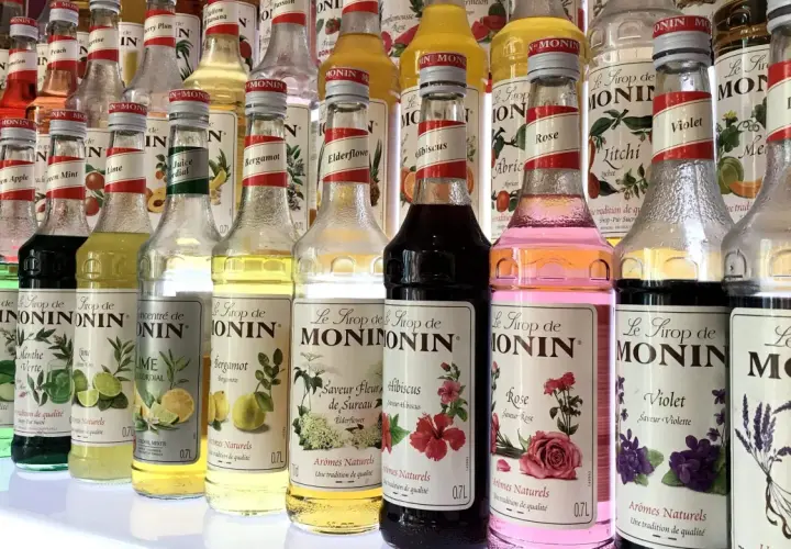 Rząd butelek syropów do kawy Monin o różnych smakach: od owocowych po kwiatowe, np. róża, lawenda, hibiskus.